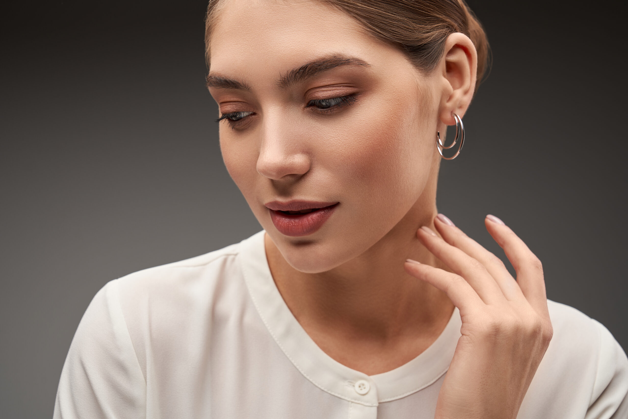 Close up portrait of young beautiful brunette female model presenting silver earrings. Front view of woman with clean skin posing, isolated on studio background. Concept of jewelry.